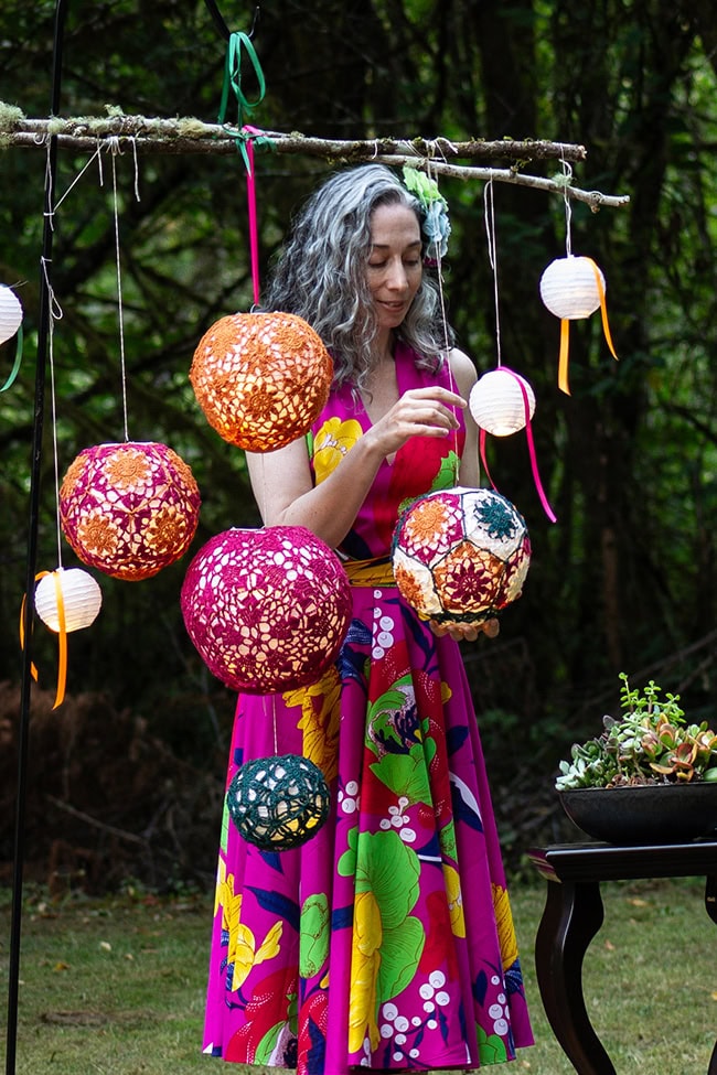 A woman standing behind the globes, holding on in her hands. The one she is holding is multicolored with the insides of each motif a different color from the outside, all joined together with teal.