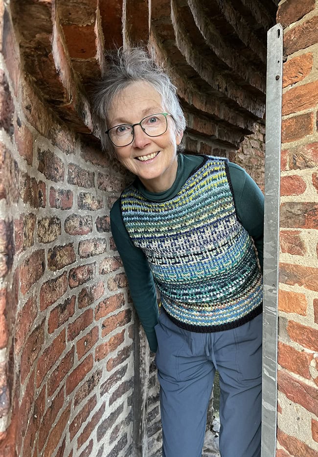 A woman peering around the corner of a brick tunnel. She is wearing a vest that is giving the impression of blues and greens that is not fair isle but is reminiscent of fair isle.