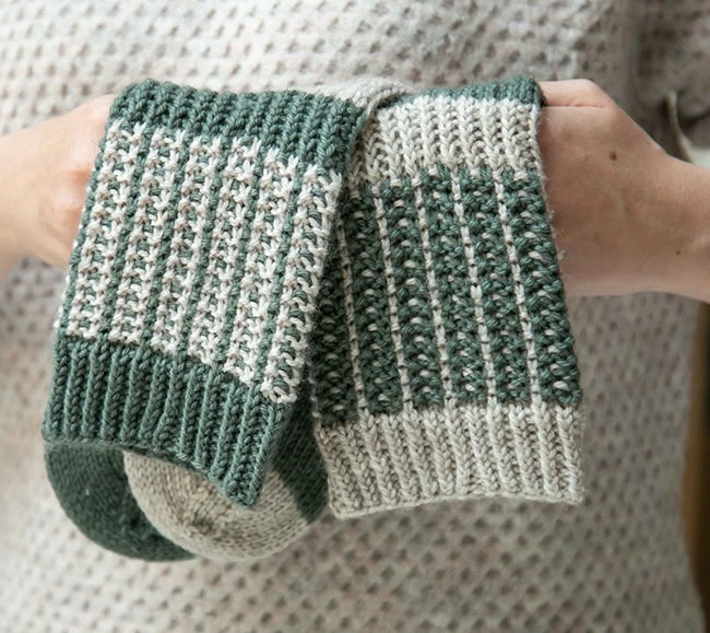 A closeup of the socks being held, showing the mosaic pattern. It looks like cream-white-cream stipes with a column of white and green alternating between them. Colors are reversed on the second sock.