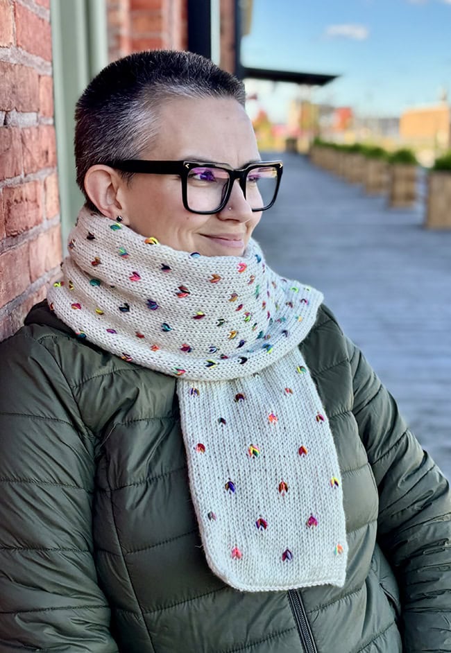 A person wearing a white scarf with multi-colored thrumbs in multiple diagonal lines across the width of the scarf.